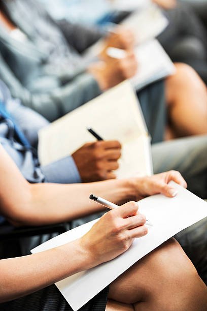 "Unrecognizable people sitting in a row holding pens, notebooks and writing. Focus on foreground."